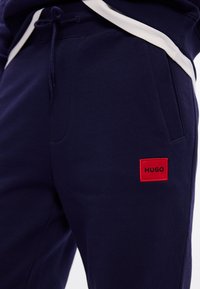 Granatowe spodnie dresowe wykonane z miękkiego materiału, z czerwonym logo HUGO w formie naszywki oraz białymi paskami po bokach. Zawierają regulowany pasek na sznurki w talii.