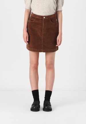 Femme portant une mini-jupe en velours côtelé marron, un pull beige, des chaussettes noires et des mocassins noirs à pampilles, debout devant un fond blanc uni.