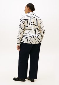 Λευκό πουκάμισο με σκούρες μπλε αφηρημένες γραμμές, μακριά μανίκια και γιακά, σε συνδυασμό με σκούρες μπλε παντελόνες με φαρδιά μπατζάκια και μαύρα παπούτσια, από πίσω όψη.