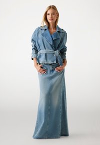 Giacca di jeans con vita con cintura e ampi risvolti, abbinata a una lunga gonna di jeans blu coordinata. Entrambi i capi presentano un lavaggio sbiadito.