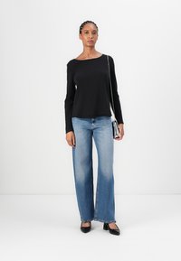 Black long-sleeve top, relaxed fit, paired with light blue wide-leg jeans. Model holds a small silver crossbody bag, wearing black pumps.
