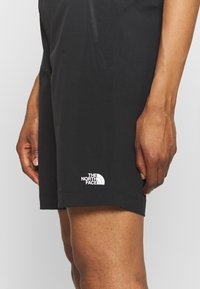 Svarta sportshorts i lätt material, med en dragkedjeficka och en vit logotyp på den vänstra nederkanten.