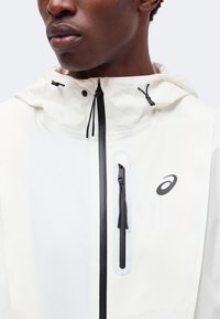 Homme noir portant une veste blanche avec une capuche, des fermetures éclair noires et un logo noir sur la poitrine, montré du cou jusqu’au milieu du torse.