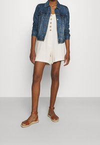 Personne portant une combinaison blanche à boutons, une veste en denim et des sandales à bride cheville marron, se tenant devant un fond uni.