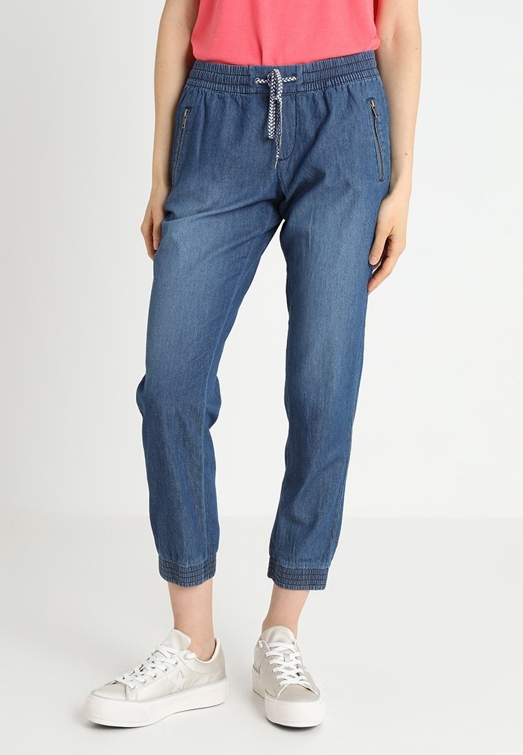 Femme portant un pantalon de jogging bleu décontracté avec une taille à cordon et des poches zippées, associée à des baskets blanches.