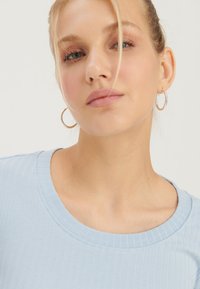 Top de culoare bleu deschis, cu un guler rotund, însoțit de cercei subțiri cu cerc. Textură netedă vizibilă pe țesătură și detalii din dantelă pe mâneci.