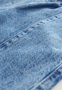 Denimtyg i mellanblått med en lätt blekning, med synlig sömnad och en texturerad yta. Närbild som betonar materialdetaljer.