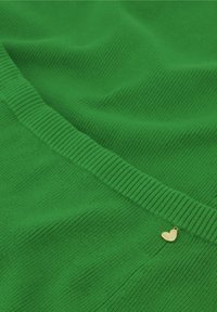 Grüner gerippter Stoff mit glatter Textur und einem subtilen Glanz. Hat eine kleine goldene herzförmige Akzentuierung am Rand.
