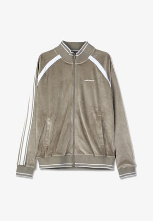 Kordfløyelsjakke med glidelås i dempet brun farge med hvite detaljer. Har en ribbet krage, sidelommer og vertikale striper på ermene.