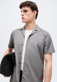 Νέος άνδρας που φοράει γκρι πουκάμισο με κοντά μανίκια πάνω από λευκό t-shirt, κρατώντας ένα διπλωμένο μαύρο σακάκι στο αριστερό του χέρι.