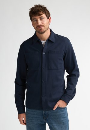 Mann mit kurzen lockigen Haaren, der ein marineblaues Hemd mit Knöpfen und zwei Brusttaschen sowie blaue Jeans trägt, steht mit einer Hand in der Tasche.