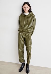 Hoodie od umjetne kože maslinasto zelene boje s vezicom i usklađenim tapered hlačama. Crne cipele sa šiljastim vrhom do gležnja upotpunjuju odjevnu kombinaciju. Glatka, sjajna tekstura.