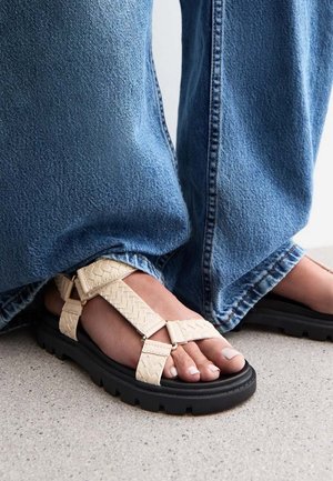 Beige woven sandals with black rubber soles. Strappy design featuring crisscross and adjustable buckle. Paired with loose blue denim.