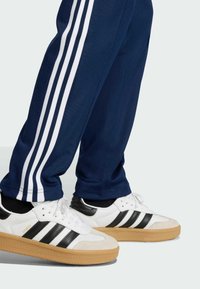 Λευκά και μαύρα αθλητικά παπούτσια με τρεις μαύρες ρίγες, σόλες από καουτσούκ καραμέλας, συνδυασμένα με ναυτικά παντελόνια αθλητικής φόρμας με τρεις λευκές ρίγες στο πλάι.