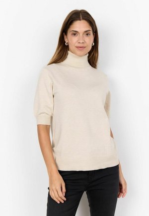 Beige lyhythihainen pooloneule, jossa on pehmeä neulostekstuuri, rento malli ja ribbauskärjet, yhdistetty mustiin housuihin.