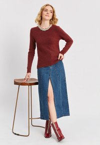 Bordo džemperis ar apaļu izgriezumu, kombinēts ar ceļa garuma džinsu svārkiem ar priekšēju šķēlumu. Modelis valkā sarkanas potīšu zābakus.