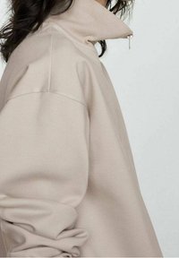 Pullover beige a collo alto con colletto a coste, dotato di dettaglio a mezza zip e vestibilità rilassata. Tessuto morbido con leggera texture.