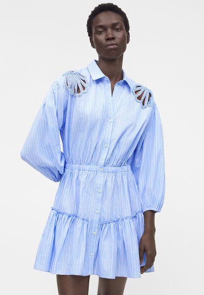 Γυναίκα που φοράει ένα ανοιχτό μπλε ριγέ φόρεμα με κουμπιά, φουσκωτά μανίκια και κομμένες λεπτομέρειες με χάντρες στους ώμους, στέκεται μπροστά σε απλό φόντο.