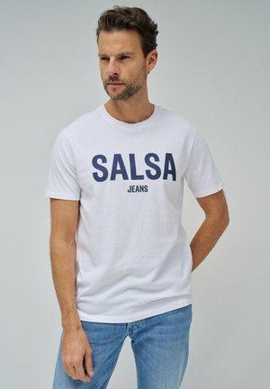 Camiseta de algodón blanca con texto en azul marino "SALSA JEANS", cuello redondo, mangas cortas y un ajuste casual, combinada con jeans de color azul claro.