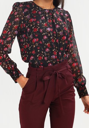 Kobieta ubrana w prześwitującą czarną bluzkę w czerwono-fioletowy kwiatowy wzór oraz bordowe spodnie z wysokim stanem, wiązane na kokardę w talii.