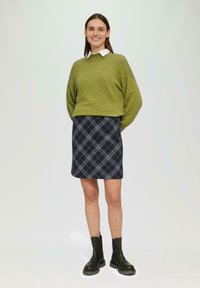 Grüner, übergroßer Pullover mit einem weißen Kragen, kombiniert mit einem marineblauen Kilt mit hellblauen Akzenten. Schwarze Knöchelstiefel runden das Outfit ab.