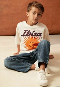 Bílý tričko s grafickým sluncem a textem "Ibiza ISLA FELIZ", doplněné o volné džíny a bílé tenisky. Neformální posed.