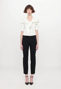 Bluză albă tricotată, cu un decolteu volanat și mâneci scurte, asortată cu pantaloni negri cu o croială slim-fit. Modelul poartă tocuri negre cu vârful ascuțit.