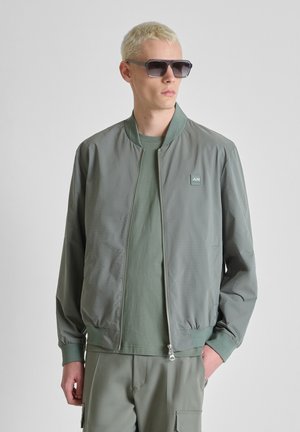 Jonge man met kort blond haar, donkere zonnebril, lichtgroene zip-up jas, bijpassend T-shirt en broek, poserend tegen een effen achtergrond.