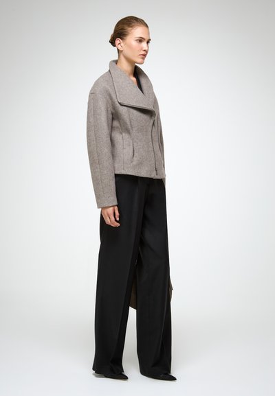 Chaqueta de mezcla de lana gris con un amplio cuello, diseño corto y cierre frontal de cremallera. Combinada con pantalones de pierna ancha negros y zapatos de punta.