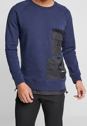 Mand iført en marineblå sweatshirt med to sorte stoflommer og en spændelås på venstre bryst, kombineret med sorte bukser.