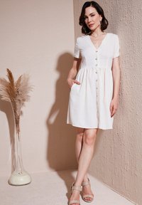 Short-sleeved, white linen dress with a V-neck, button front, and pockets. The skirt is slightly flared, creating a relaxed silhouette.