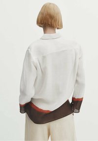 White linen shirt with a curved hem, featuring a brown and red accent at the bottom. Long sleeves and a classic collar detail.