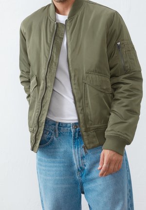Hombre con chaqueta bomber verde oliva sin cerrar sobre camiseta blanca y vaqueros azules, con las manos relajadas a los lados.