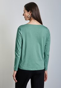 Pitkähihaista toppia pehmeästä, teal-värisestä puuvillasta, jossa on pyöreä kaula-aukko ja normaali istuvuus. Ominaisuuksina sileä rakenne ja yksinkertainen helma. Yhdistetty mustiin housuihin.