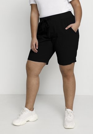 Pantalones cortos negros con una cintura ajustable, que cuentan con bolsillos laterales y dobladillos enrollados. Llevados con zapatillas de color claro.