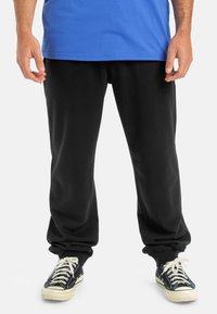 Pantalones deportivos negros con corte relajado, puños acanalados y bolsillos laterales, combinados con una camiseta azul y zapatillas negras. Material de mezcla de algodón suave.