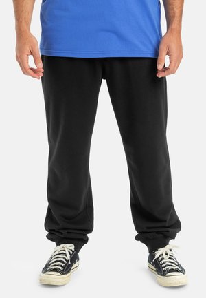 Pantalones deportivos negros con corte relajado, puños acanalados y bolsillos laterales, combinados con una camiseta azul y zapatillas negras. Material de mezcla de algodón suave.