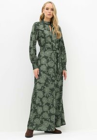 Lange groene bloemenjurk met lange mouwen, gerimpelde kraag, strik om de taille en een ingewikkeld bloemenpatroon. Zachte stof met een gladde textuur.