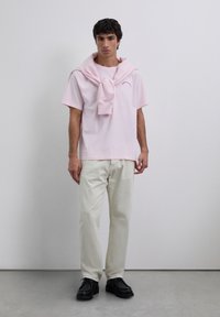 Mężczyzna w jasnoróżowej koszulce z pasującym swetrem założonym na ramionach, w off-white spodniach i czarnych butach, stojący przed gładką ścianą.