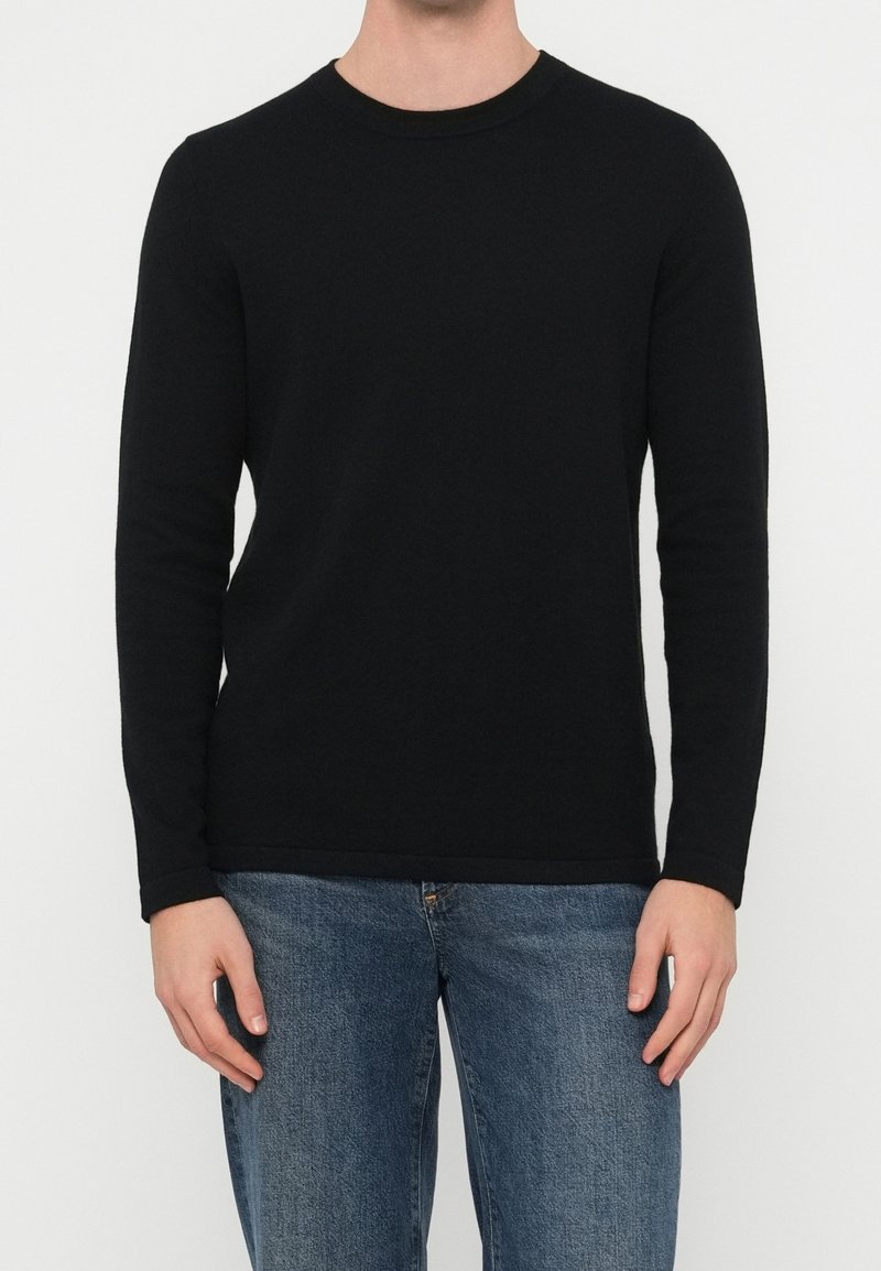 Homme portant un t-shirt noir uni à manches longues et un jean bleu, debout devant un fond blanc, les bras détendus le long du corps.