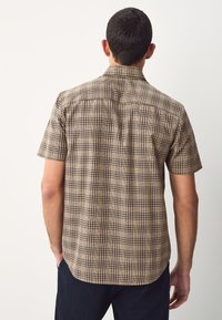 Man facing away wearing short-sleeve beige and brown plaid shirt and dark pants against a plain light background.