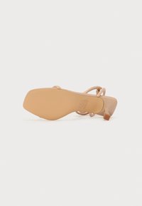 Sandale à talon aiguille beige avec un bout carré, un design à brides croisées et un upper en synthétique lisse. Semelle en caoutchouc texturé avec un détail de marque.