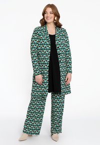 Muster langer Mantel und passende weite Hose in Grün, Creme und Rosttönen, mit einem abstrakten geometrischen Design. Schwarzes Top darunter.