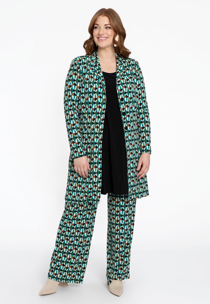 Muster langer Mantel und passende weite Hose in Grün, Creme und Rosttönen, mit einem abstrakten geometrischen Design. Schwarzes Top darunter.