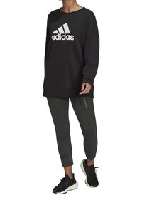 Sudadera negra con el logo blanco de "adidas", combinada con pantalones grises ajustados que tienen tres rayas verticales y zapatillas deportivas negras.