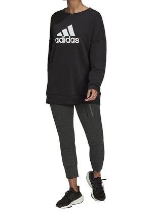 Sweatshirt noir avec le logo "adidas" blanc, associé à un pantalon gris fuselé avec trois bandes verticales et des chaussures de sport noires.