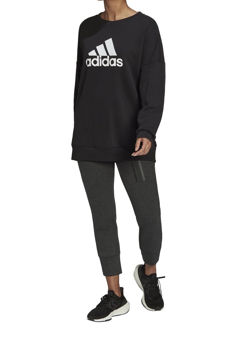 Sudadera negra con el logo blanco de "adidas", combinada con pantalones grises ajustados que tienen tres rayas verticales y zapatillas deportivas negras.
