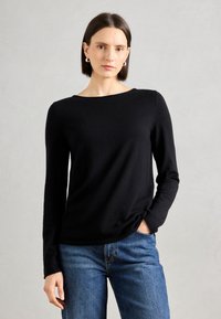 Schwarzes Langarmoberteil mit Bootsausschnitt, weicher Textur und lockerer Passform. Getragen mit blauer Jeans, die am Saum einen leichten Twist hat.