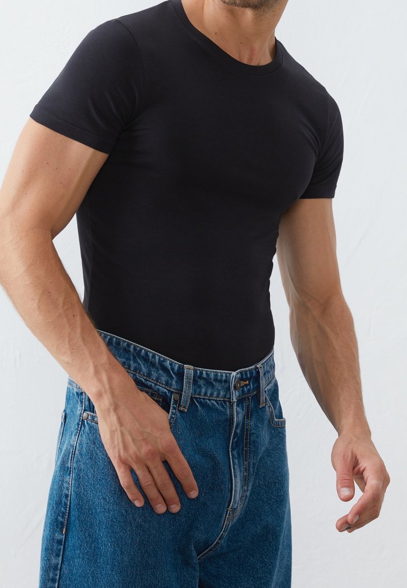 Homme portant une chemise noire cintrée à manches courtes rentrée dans un jean bleu taille haute sur fond uni.
