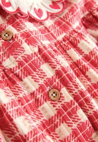 Rochie cu un model roșu și crem, având un motif de frunze, confecționată dintr-un material moale și cu nasturi din lemn pentru închidere. Detaliu cu decolteu ondulat.